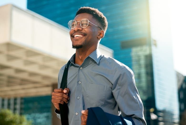 Smiling Man with Glasses Walking Downtown – Optometrist Houston Confident man wearing stylish round glasses and smiling while walking through a sunny cityscape, representing clear vision and confidence. – Optometrist Houston