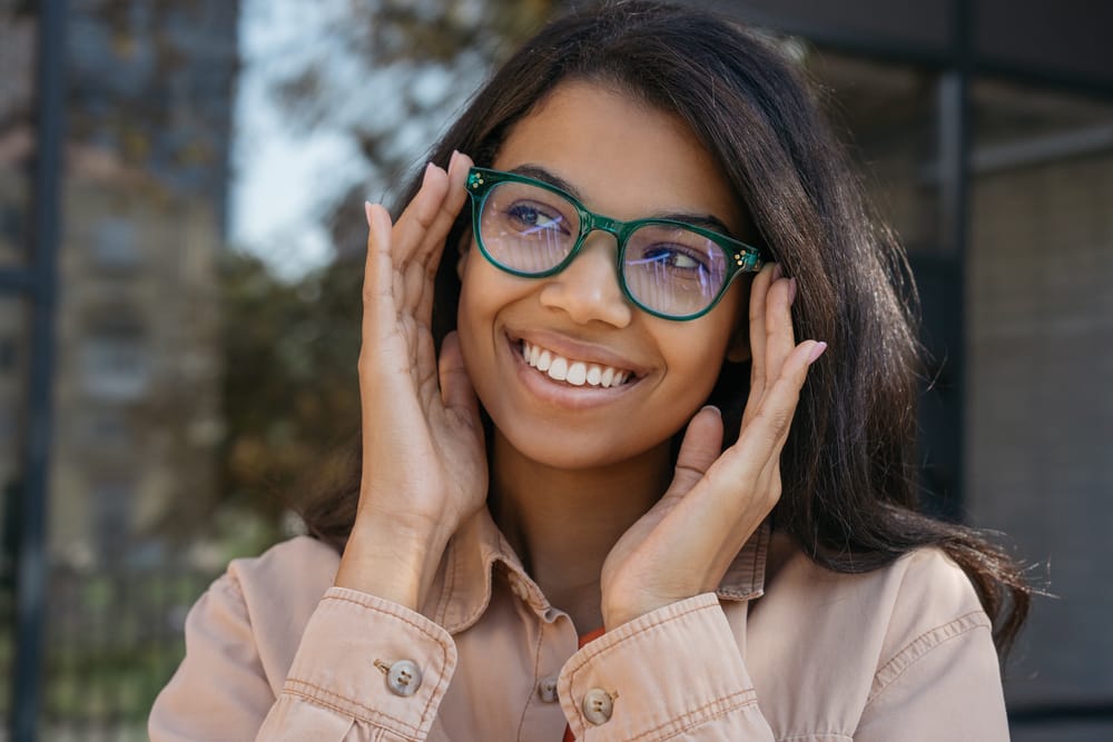 Smiling Woman Wearing Green Prescription Glasses Outdoors – Eyeglasses Houston TX Young woman with long dark hair wearing stylish green eyeglasses and a beige shirt, smiling confidently outdoors in natural light – Eyeglasses Houston TX
