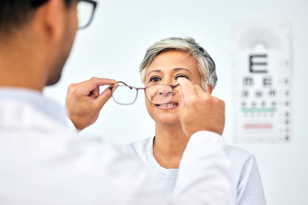 Optometrist Adjusting Eyeglasses for Mature Patient During Vision Check – Houston Eye Exam Optometrist fitting eyeglasses on a smiling woman with short gray hair during a vision test, with an eye chart in the background. – Houston Eye Exam
