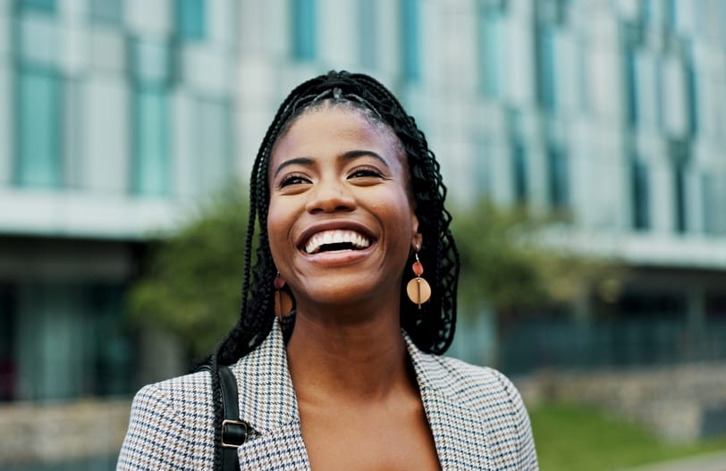 Joyful woman with braids smiling brightly while standing outdoors near a modern office building, symbolizing clear vision and confidence. – Bellaire Eye Doctor