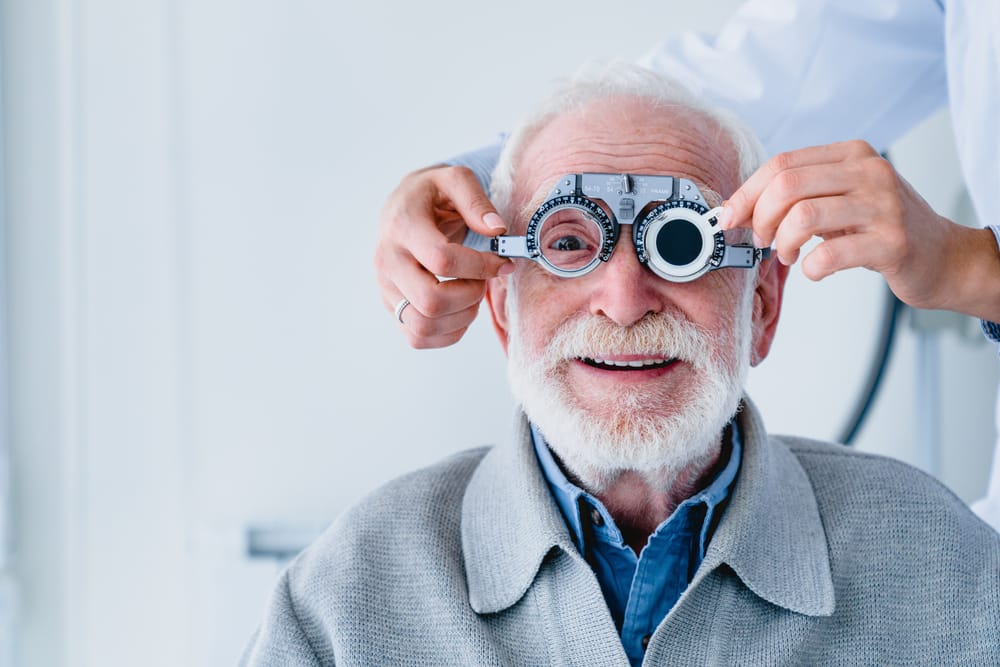 Senior Man Undergoing Vision Test with Phoropter Device – Houston Eye Exam Elderly man smiling while an optometrist adjusts a phoropter device during an eye examination at a clinic. – Houston Eye Exam