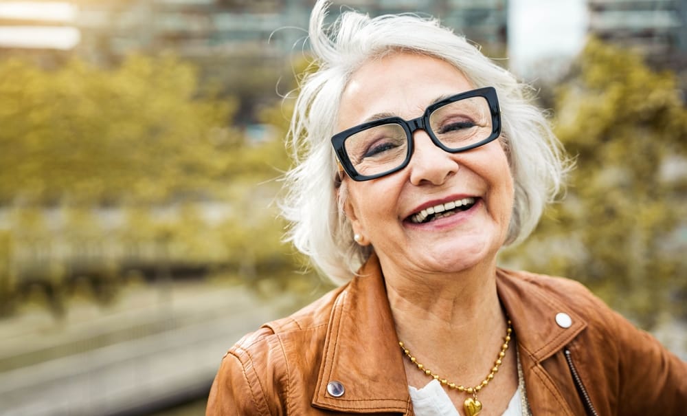 Stylish Senior Woman Smiling in Trendy Eyeglasses – Eyeglasses Houston TX Joyful older woman with short gray hair wearing modern black eyeglasses and a brown leather jacket, symbolizing style and confidence with quality eyewear – Eyeglasses Houston TX