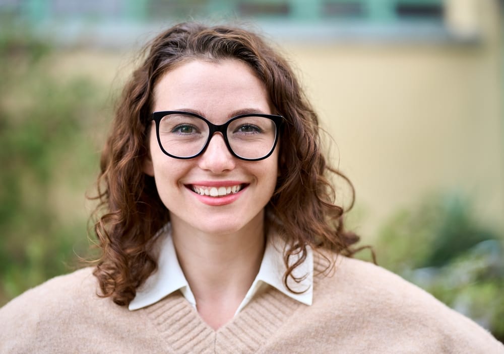 Smiling Woman Wearing Stylish Prescription Glasses – Eyeglasses Houston TX Happy woman with curly hair and fashionable black eyeglasses smiling outdoors, representing stylish and comfortable eyewear options – Eyeglasses Houston TX