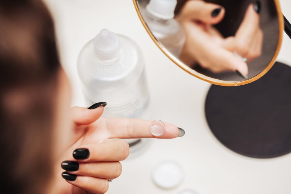 Contact Lens on Finger – Eye Doctor Contacts A close-up of a hand with black nail polish holding a contact lens on the fingertip next to a mirror, contact lens case, and solution bottle – Eye Doctor Contacts