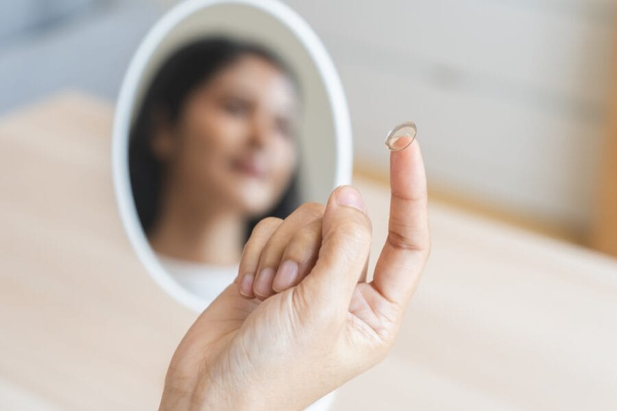 A person holding a contact lens on their fingertip with a blurred reflection of their face in a mirror – Can You Wear Contact Lenses if You Need Bifocals