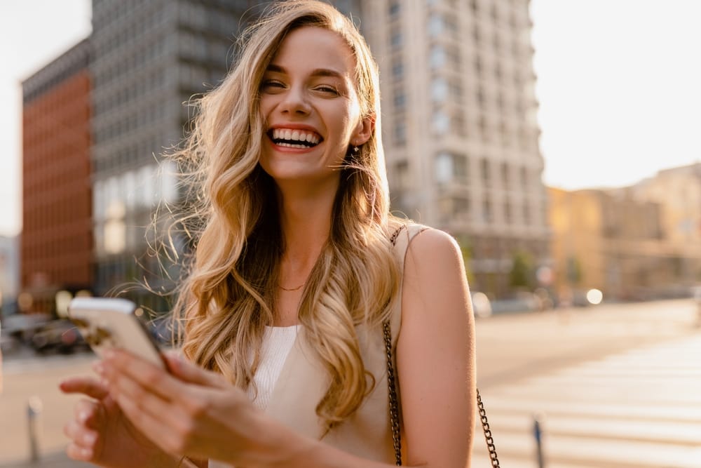 Smiling Woman with Long Blonde Hair Holding a Phone Outdoors – Vision Corner Cheerful woman with long blonde hair smiling brightly while holding her phone in the city, showcasing clear and confident vision. – Vision Corner